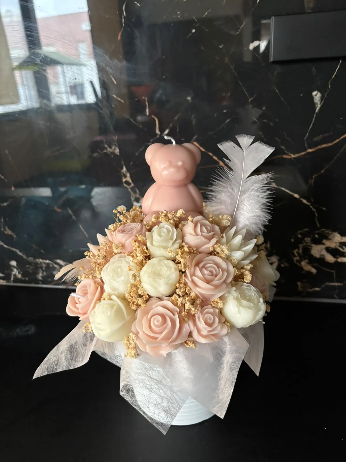 Bouquet de rosas de cera rosa e creme com uma vela em forma de ursinho cor-de-rosa e uma pena branca, num vaso embrulhado em tule branco
