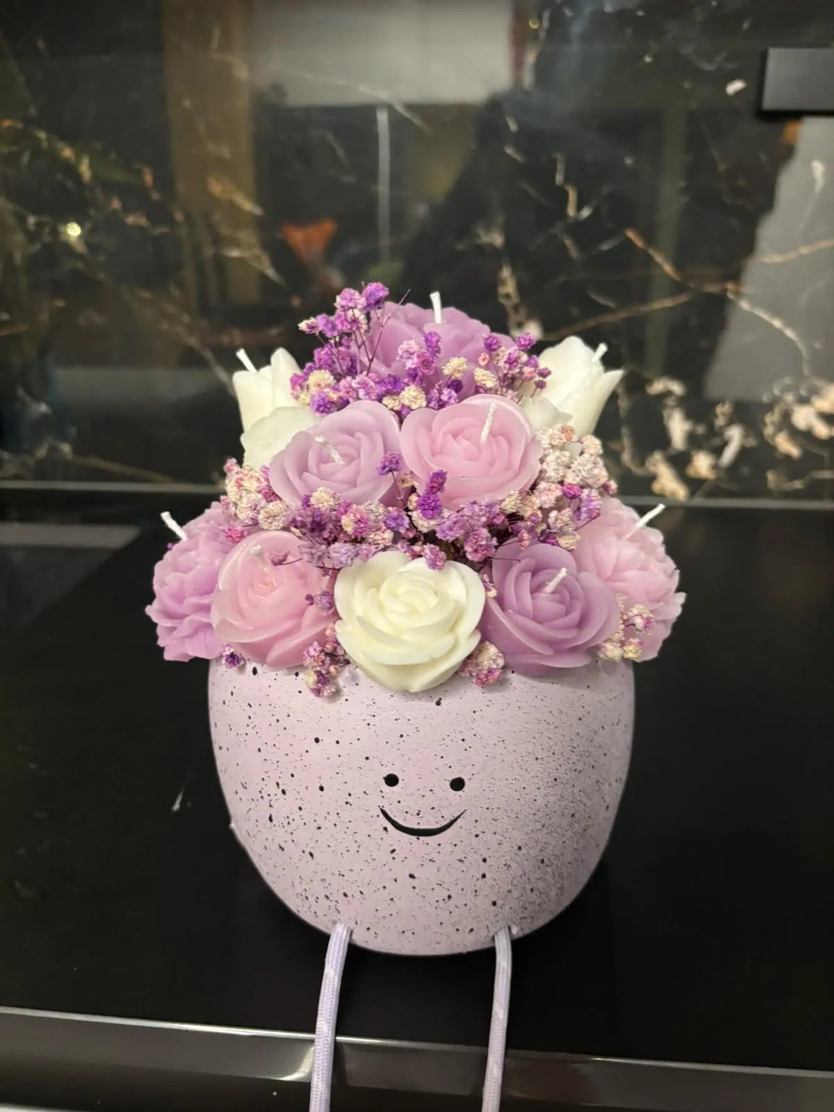 Bouquet de rosas de cera lilás, rosa e creme num vaso cor-de-rosa salpicado com uma cara sorridente desenhada e duas perninhas penduradas